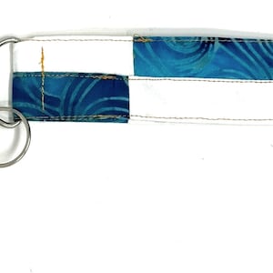 Pode incluir: Um chaveiro de tecido com padrão branco e azul, com um anel de chave prateado e um pequeno tassel laranja.