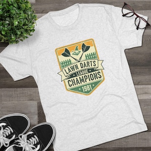 May include: A light grey t-shirt with a retro graphic design. The design features the text "Lawn Darts League Champions 1981", lawn darts, and a fence. Black trainers and spectacles are also visible in the image.