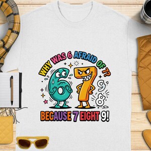 Könnte beinhalten: Weißes T-Shirt mit einer bunten Cartoon-Grafik mit den Zahlen 6, 7, 8 und 9. Der Text auf dem Shirt lautet: „WARUM HATTE 6 ANGST VOR 7? WEIL 7 ACHT 9!“ Ein lustiges, verspieltes Design.