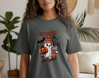 May include: A dark grey t-shirt with a Halloween design. The graphic includes a ghost in an orange beanie, holding a pumpkin and a drink. The words "spooky season" are written above and below the image in orange script.