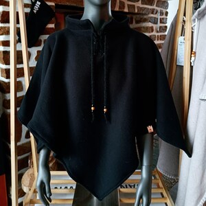 May include: A black poncho with a high collar and a drawstring closure. The poncho has a unique, asymmetrical design and is displayed on a mannequin. The drawstring features wooden beads. The overall style is casual and practical.