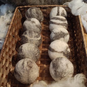 Könnte beinhalten: Ein Weidenkorb gefüllt mit grauen und weißen Woll-Trocknerbällen. Die Bälle sind in Reihen angeordnet, einige zeigen ein gestreiftes Muster. Flauschige weiße und graue Wollfasern umgeben den Korb.