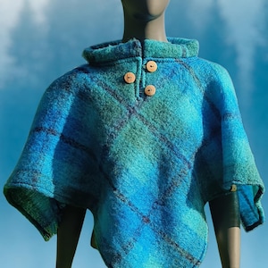 Puede incluir: Un poncho de cuadros en tonos azul y verde azulado con cuello alto y tres botones de madera de color marrón claro en el escote. La prenda tiene una superficie texturizada y un dobladillo en punta, exhibida sobre un maniquí.