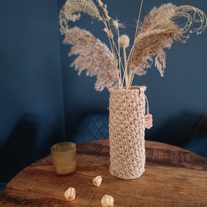 Może przedstawiać: Cylindryczny wazon z plecionej juty, wypełniony suszonymi trawami i główkami nasion. Wazon stoi na drewnianym stole. Na stole znajduje się również mały szklany świecznik i trzy suszone strąki nasion.