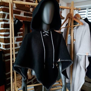Peut inclure: Une cape noire à capuche avec des coutures blanches sur les bords et un cordon de serrage blanc. La cape est présentée sur un mannequin. D'autres capes sont visibles en arrière-plan.