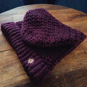 May include: A knitted burgundy hat and matching cowl scarf set. The set is made of a thick, textured yarn and features a decorative button on the cowl. The items are displayed on a wooden surface.