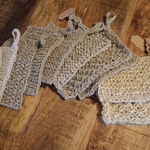 Peut inclure: Plusieurs gants de toilette en jute beige avec des poignées en boucle, disposés sur une surface en bois. Les gants de toilette sont rectangulaires avec des coins arrondis et ont un tissage texturé. Certains ont de petites étiquettes attachées.