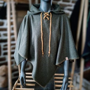 May include: A dark gray hooded poncho with a tan drawstring closure. The poncho has a unique asymmetrical hemline and tan stitching along the edges. The garment is displayed on a mannequin.