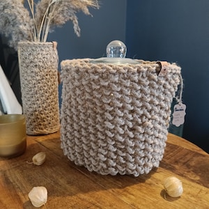 Peut inclure: Un pot beige tricoté à la main avec un couvercle en verre, à côté d'un vase assorti avec des plantes séchées. Le pot a un aspect texturé et tissé et une petite étiquette. Les articles sont sur une surface en bois.