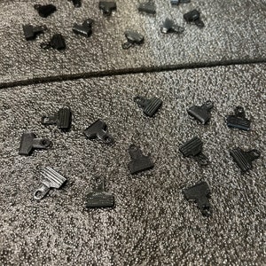 May include: A collection of black plastic clips scattered on a gray textured surface. The clips are small and have a simple design.
