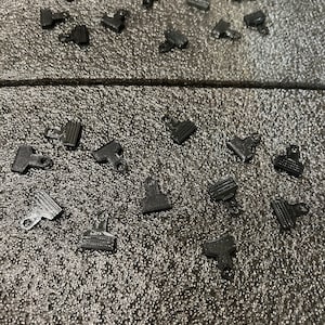 May include: A collection of black plastic clips scattered on a gray textured surface. The clips are small and have a simple design.
