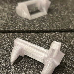 May include: Two white plastic 3D printed objects. The first object is a rectangular frame with a square opening. The second object is a U-shaped frame with two pointed ends.