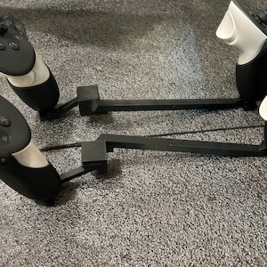 May include: Two black VR controllers with white faceplates are mounted on black adjustable arms. The arms are attached to a grey surface.