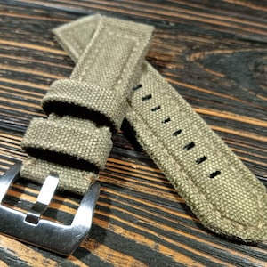 May include: A khaki-colored canvas watch strap with a stainless steel buckle. The strap has multiple adjustment holes and a textured weave. The buckle is silver and rectangular. The watch strap is designed to fit a standard watch.