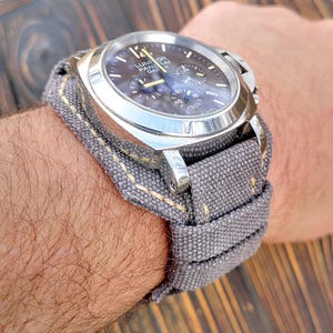Op de afbeelding: Een zilveren chronograafhorloge met een grijze canvas horlogeband. De wijzerplaat van het horloge heeft een zwarte achtergrond met witte markeringen en de tekst "Luminor Panerai Day Date" erop gedrukt.