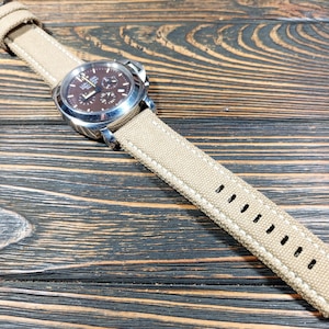 Op de afbeelding: Een polshorloge met een bruine wijzerplaat en een beige canvas band. Het horloge heeft een zilverkleurige lunette en een chronograaf ontwerp. De band is gestikt met witte draad en heeft verstelgaten. Het horloge ligt op een donkere houtnerf achtergrond.