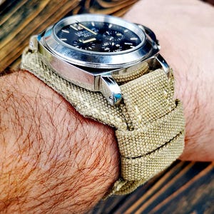 May include: A silver wristwatch with a black face and a beige canvas watch strap. The watch face has the text "L.U.T.E.C. PATEK PHILIPPE GENEVE" on it.