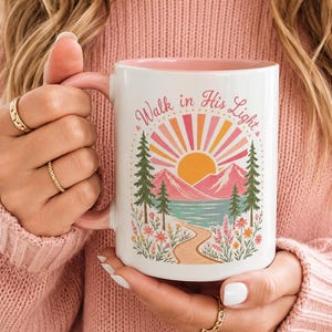 Può includere: Una tazza in ceramica bianca con interno rosa che presenta una grafica colorata di un sole, montagne, pini e un sentiero. Il testo "Walk in His Light" è stampato in un arco curvo sopra il sole con un carattere tipografico rosa.
