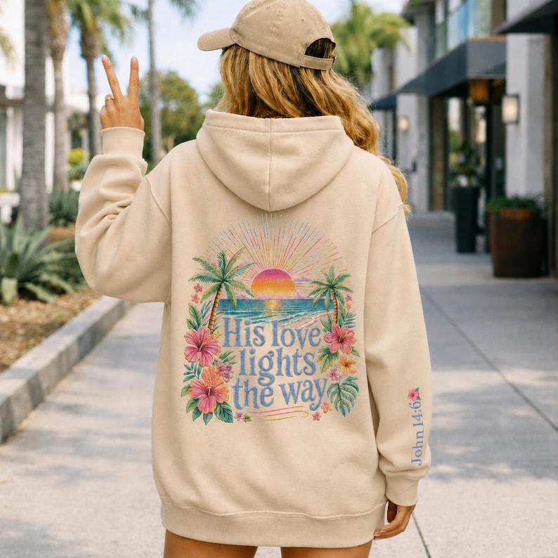 May include: A beige hooded sweatshirt featuring a back graphic of a sunset over the ocean, palm trees, and hibiscus flowers. The text reads, "His love lights the way." The sleeve includes the text "John 14:6" with a small flower icon.