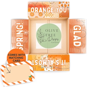 May include: An open cardboard box with orange-themed patterns and the text ORANGE YOU GLAD IT'S ALMOST SPRING! A circular green sticker in the center reads OLIVE TREE DESIGNS by jacqueline. A matching rectangular card with orange stripes is included.