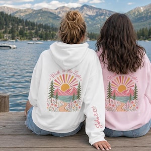 May include: A white hooded sweatshirt featuring a back graphic of a sun, mountains, pine trees, and a path with the text Walk in His Light and John 8:12 on the sleeve. A light pink hooded sweatshirt with an identical graphic is also visible.