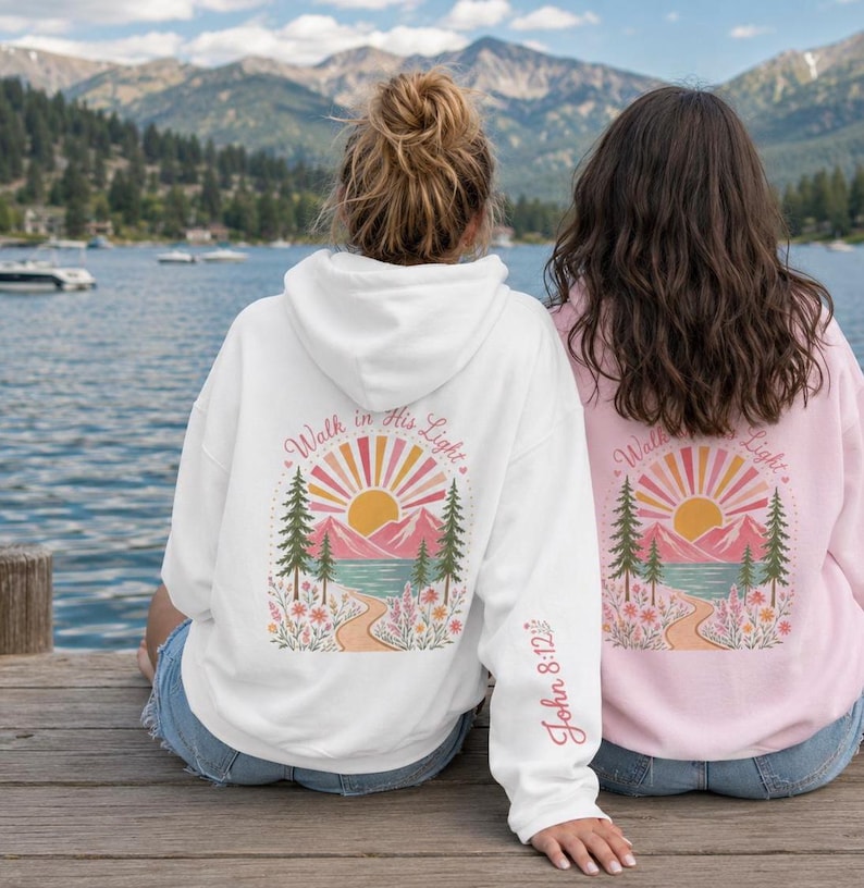 May include: A white hooded sweatshirt featuring a back graphic of a sun, mountains, pine trees, and a path with the text Walk in His Light and John 8:12 on the sleeve. A light pink hooded sweatshirt with an identical graphic is also visible.
