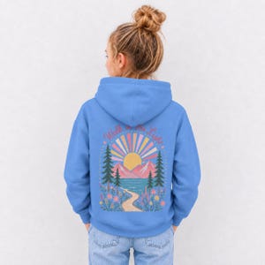 May include: A bright blue hooded sweatshirt featuring a back graphic of a yellow sun, pink mountains, a blue lake, and pine trees. A winding path leads through colorful flowers. The text "Walk in the Light" is arched above the sun design.
