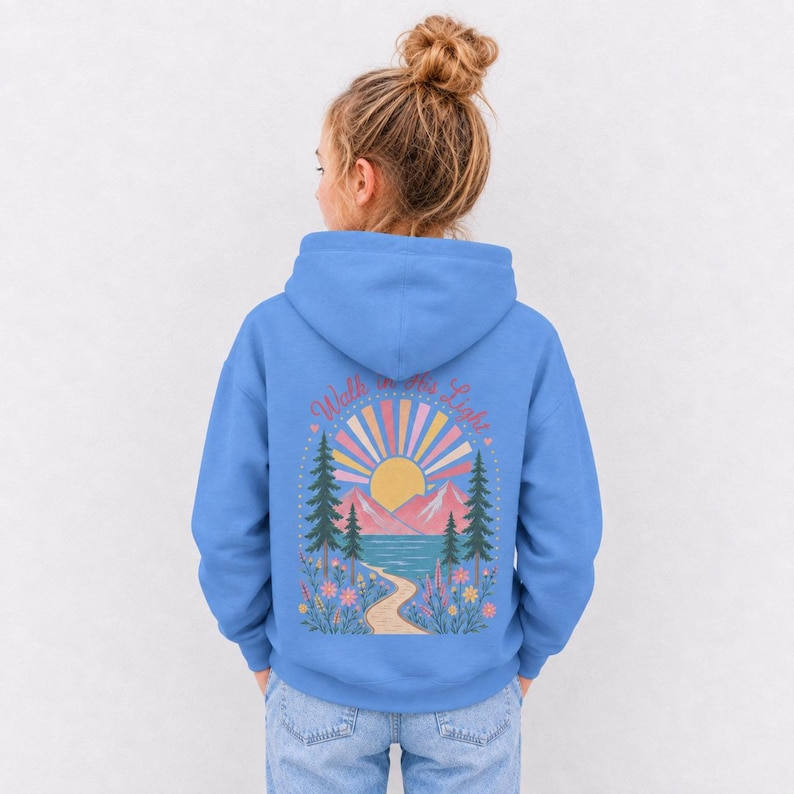 May include: A bright blue hooded sweatshirt featuring a back graphic of a yellow sun, pink mountains, a blue lake, and pine trees. A winding path leads through colorful flowers. The text "Walk in the Light" is arched above the sun design.