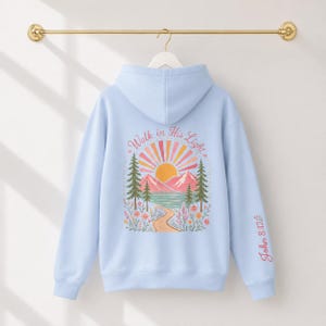 A light blue Christian hoodie featuring a mountain lake graphic on the back. The design displays a sun, mountains, pine trees, and a path with the text Walk in His Light surrounded by colorful flowers in shades of pink, orange, and green.