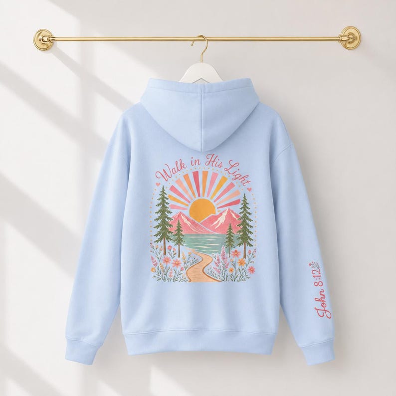 A light blue Christian hoodie featuring a mountain lake graphic on the back. The design displays a sun, mountains, pine trees, and a path with the text Walk in His Light surrounded by colorful flowers in shades of pink, orange, and green.