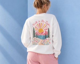 May include: A white long-sleeve sweatshirt featuring a back graphic of a sun, mountains, pine trees, and a path with the text Walk in His Light. The design includes pink, yellow, blue, and green colors. The sweatshirt is paired with pink pants.
