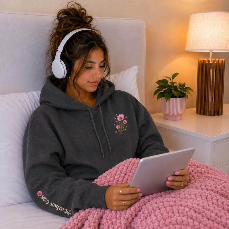 May include: A dark charcoal gray hooded sweatshirt featuring a small pink flower design on the chest and the text Matthew 6:28 printed on the sleeve. The hoodie has long sleeves, a drawstring hood, and a front kangaroo pocket.