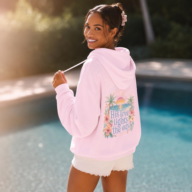 May include: A pink hooded sweatshirt featuring a small graphic on the upper left chest. The graphic depicts a tropical scene with a palm tree, a pink and orange sunset, and blue ocean waves. The hoodie includes long sleeves and a front pouch pocket.