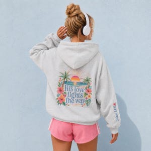 May include: A light gray hooded sweatshirt featuring a back graphic of a tropical beach scene with palm trees, a sunset, and hibiscus flowers. The text "His love lights the way" is printed in blue, and "John 14:6" appears on the sleeve.