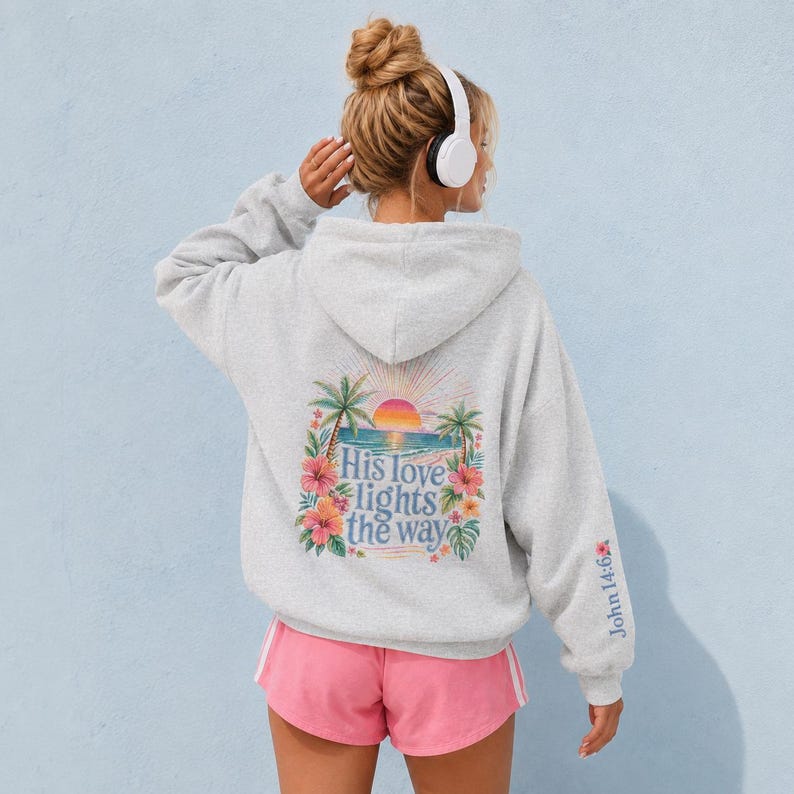 May include: A light gray hooded sweatshirt featuring a back graphic of a tropical beach scene with palm trees, a sunset, and hibiscus flowers. The text "His love lights the way" is printed in blue, and "John 14:6" appears on the sleeve.