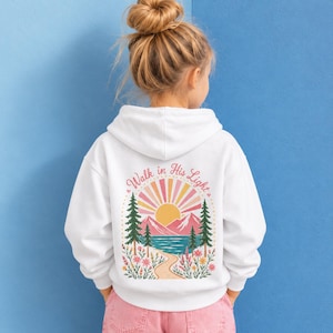 May include: A white hooded sweatshirt featuring a back graphic of a sun, mountains, lake, pine trees, and flowers. The text "Walk in His Light" is printed in a curved arch above the sun, which is colored in shades of pink, yellow, and orange.