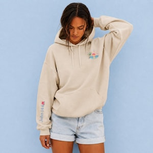 May include: A beige hooded sweatshirt featuring a small graphic on the upper left chest. The graphic depicts a tropical scene with a palm tree, a pink and orange sunset, and blue ocean waves. The hoodie includes long sleeves and a front pouch pocket.