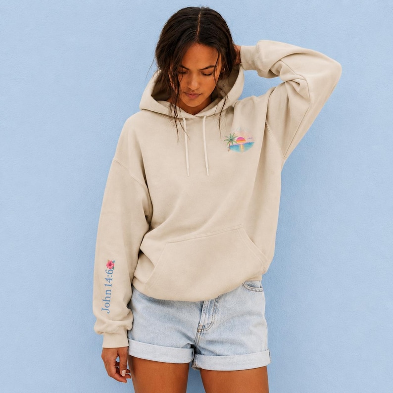 May include: A beige hooded sweatshirt featuring a small graphic on the upper left chest. The graphic depicts a tropical scene with a palm tree, a pink and orange sunset, and blue ocean waves. The hoodie includes long sleeves and a front pouch pocket.