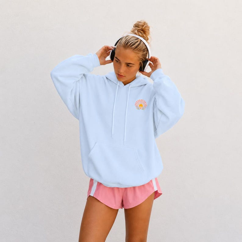 A light blue hooded sweatshirt featuring a small graphic of a sun with rays on the left chest, paired with pink athletic shorts. The hoodie includes a front kangaroo pocket and matching light blue drawstrings at the neckline. Christian hoodie