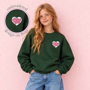 May include: A dark green long-sleeve sweatshirt featuring a white heart-shaped patch on the left chest. The patch contains the pink embroidered text "Jesus loves you!" in a retro font. The sweatshirt has a crew neckline and ribbed cuffs.