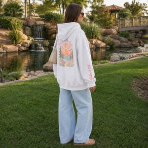 May include: A light gray hooded sweatshirt featuring a colorful graphic on the back with a sun, mountains, trees, and flowers, along with the text "Walk in His Light". The sleeve includes the text "John 8:12" in pink, paired with light blue jeans.