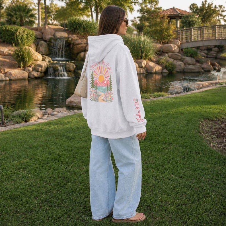 May include: A light gray hooded sweatshirt featuring a colorful graphic on the back with a sun, mountains, trees, and flowers, along with the text "Walk in His Light". The sleeve includes the text "John 8:12" in pink, paired with light blue jeans.