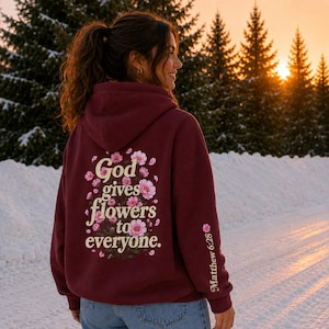 May include: A maroon hooded sweatshirt featuring a back graphic of pink flowers and the text "God gives flowers to everyone." The sleeve includes a small pink flower icon and the text "Matthew 6:28" printed in a light cream color.