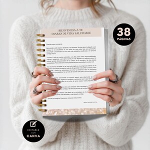 May include: A spiral-bound notebook with gold-colored coils is held open, displaying text in Spanish. The cover reads "BIENVENIDA A TU DIARIO DE VIDA SALUDABLE." A black circle with "38 P&Aacute;GINAS" is visible. The notebook is "EDITABLE CON CANVA."