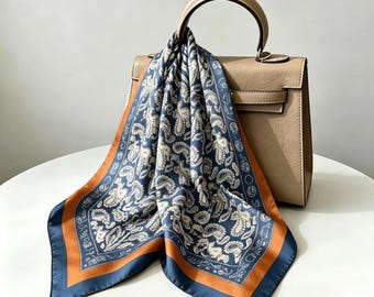 Puede incluir: Un pañuelo cuadrado de seda con un estampado de cachemira en azul y crema con un borde naranja sólido, drapeado sobre un bolso beige estructurado con un asa superior y cierre de solapa con un detalle de hebilla metálica.