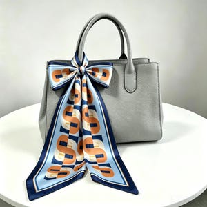 May include: A light gray structured handbag with two top handles, featuring a decorative silk scarf tied in a bow around one handle. The scarf has a light blue background with a repeating pattern of large orange letters and dark blue borders.