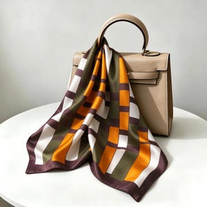 May include: A beige structured handbag with a single top handle, draped with a square silk scarf featuring a geometric pattern of intersecting blocks in shades of olive green, deep brown, bright orange, and white, creating a bold grid design.