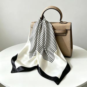 Op de afbeelding: Een gestructureerde beige handtas met een enkel handvat, gedrapeerd met een vierkante zijden sjaal. De sjaal heeft een witte basis met zwarte geometrische lijnen die een chevronpatroon vormen en een effen zwarte rand aan de onderkant.