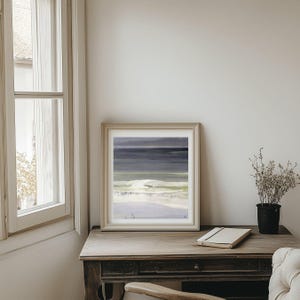 Puede incluir: Impresión artística enmarcada de un paisaje abstracto. La obra presenta una serena escena costera con tonos de azul, gris y blanco. La impresión se exhibe sobre una mesa de madera, junto a un cuaderno y una pequeña planta en maceta.