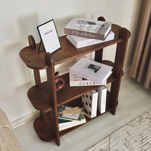 May include: A dark brown wooden bookshelf with three oval shelves. The shelves hold books, decorative boxes, and a small vase. A framed print with the text "ENGLISH HOME" is on the top shelf. The bookshelf is against a light-colored wall.